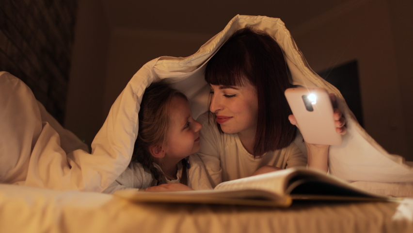 Smiling young mommy lying in bed under a blanket with little cute daughter, reading fairytales in the evening bedroom. Adorable preschool kid girl and lovely mom enjoying bedtime, touching noses | Shutterstock HD Video #1088135343