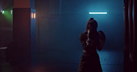 Young sports woman professionally beats a punching bag in a dark gym, smokes. Young sportswoman strikes a punching bag, professional training. Sportswoman silhouette. 4k ProRes