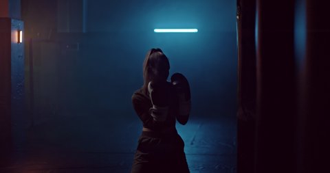Silhouette, a woman professionally boxing a punching bag in a dark gym, smoking. Woman trains punches in boxing gloves, preparing for a fight. Young woman athlete in training. 4k, ProRes