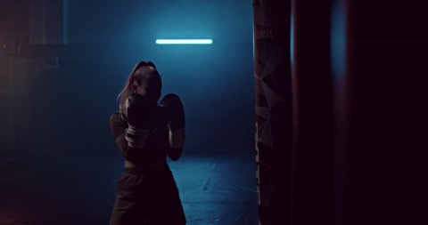 Silhouette, a young sportswoman beats a punching bag in a dark gym, smoke tubers. Professional athlete practice punches before a fight in a fight club. 4k ProRes