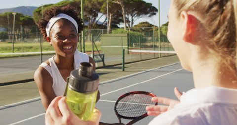 Video of happy diverse female tennis players talking and drinking water. professional tennis training, sport and competition concept.