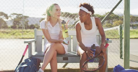 Video of happy diverse female tennis players sitting, talking and resting after match. professional tennis training, sport and competition concept.