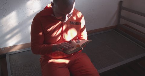 Reading in the prison. prisoner in orange uniform reading a book in a prison cell. a prisoner reads a book of prayers and looks out the window through the bars. religion in prison