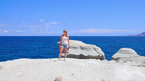 Pretty woman on the edge of the Sarakiniko cliffs with ocean in the background turns to face the camera in slow motion