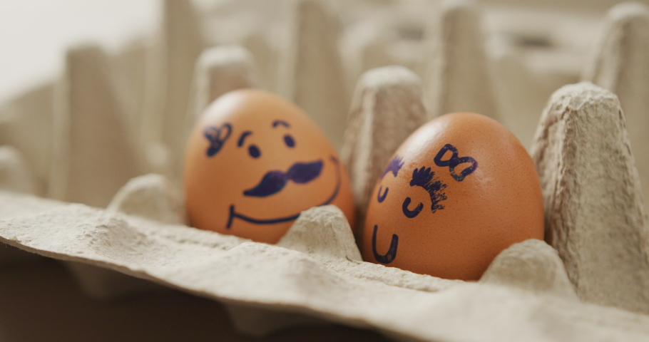Video of close up of two brown eggs with drawn faces in egg carton background. fusion food, baking, eggs and easter concept.