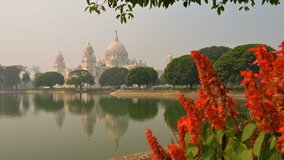 4K stock video of Victoria Memorial, a large marble building in Central Kolkata, West Bengal, India. Reflection on the lake water in midddleground. Red flowers on the right side of the frame.