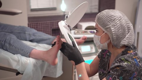 The pedicure master looks at the patient's foot through a large magnifying glass. Procedure at the podiatrist in the salon.