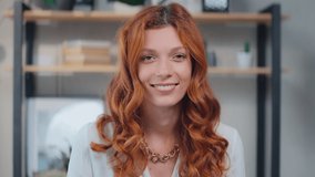 Young beautiful redhead Caucasian woman looking at camera cutely blinking and smiling listens to interlocutor during video call standing on background home interior. Headshot screen view