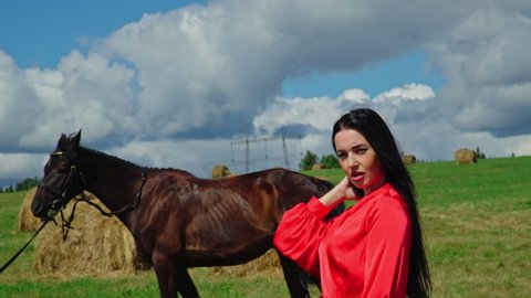 Attractive Girl in red dress straightens her hair in windy weather. Girl straightens her hair in windy weather. Girl on the background of a dark horse and hay