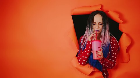 Barmaid with drink looking through hole in paper background. Female bartender with dreadlocks and cup of sweet beverage peeking through ripped orange paper background in studio and looking at camera