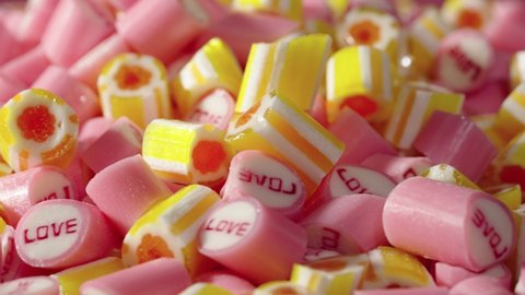 Colored striped caramel candies with inscription Love, rotating shot. Mix of pink and yellow lollipops close up. Sweet sugar dessert. Candy Shop. Festive background for holiday or children's birthday.
