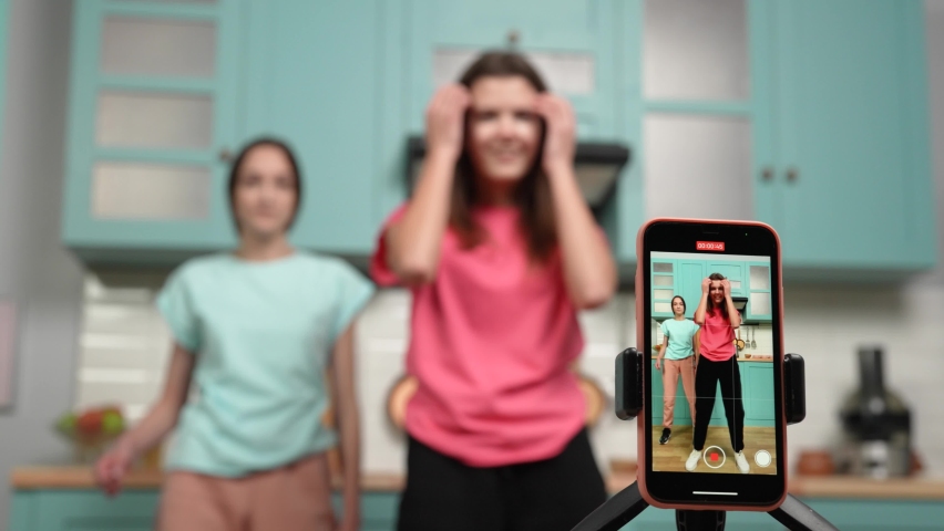 Focus on phone screen with two young girls dancing and making videos for their blogs or social networks. Bloggers are recording vlog with trend content in kitchen. Slow motion ready, 4K at 59.97fps.