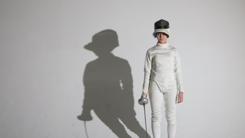Portrait of a young woman fencer saluting with rapier, putting on helmet and throwing attacking blows. An athlete poses in dark studio on white background with shadow. Slow motion. Close up.
