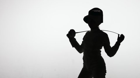 Silhouette of a young female fencer removing a protective helmet and holding a sword behind her head. Sportswoman in a white uniform poses in a dark studio on white background. Slow motion.