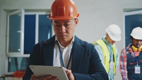 Young asian engineer, architect wearing a white protective helmet in a suit use tablet computer stand look around. On bakground builders in hardhats working in building. Slow motion