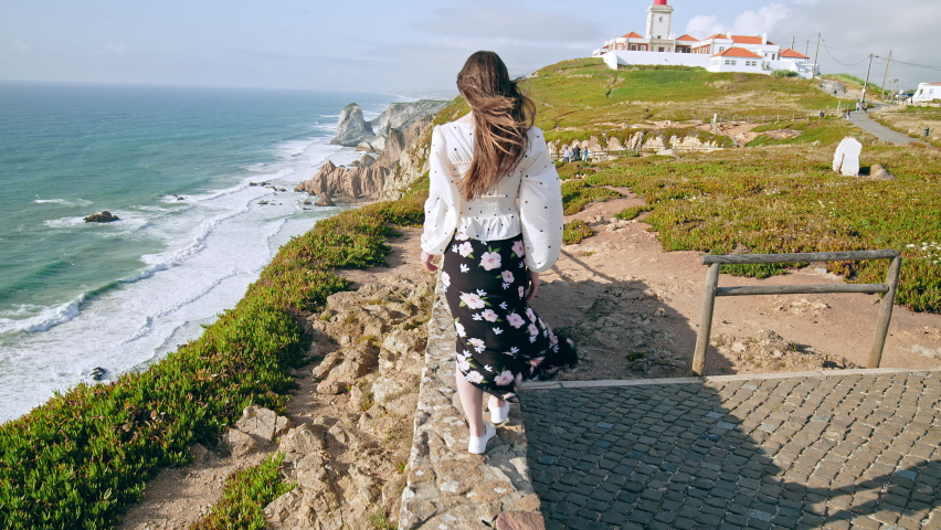 Beautiful Fashion Girl Walking in Slow Motion by the edge of a rocky cliff. Wind blows airy hair shining in the sun of a young woman in a dress walking by Farol do Cabo de São Vicente Lighthouse.