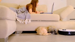 Young woman watches video via laptop small dog lies near robot vacuum cleaner on floor in living room. Appliance for comfortable life