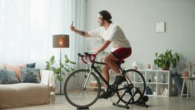 Sportsman riding bicycle and talking on video call, home gym. Young man cycling in living room with virtual conference, exercising in morning. Spanish guy working out on stationary bike.