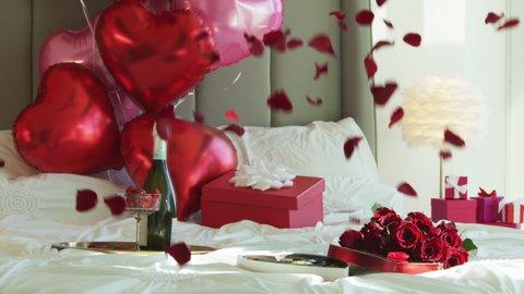 Super slow motion of falling red rose petals on white bed in elegant luxury hotel room with champaign red heart shaped balloons on background. Cinematic romantic background shot on RED camera 6K