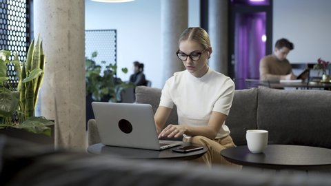 Creative industry worker is sitting in modern office or co-working hub space and manage his business with laptop computer. She finishes her task, turn off the laptop and walk away from the frame.