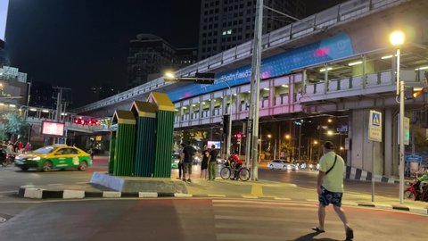 BANGKOK, THAILAND - Circa February, 2022: Timelapse 4K people at zebra crossing walking and riding scooter. Cars, motorcycle, tuk tuk at intersection and BTS skytrain transit above near Terminal 21