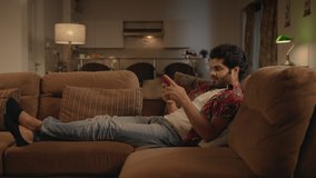 A young modern Asian Indian man or male is resting on a couch on a mobile phone or smartphone to use social media or type a text message in an interior night house setup. Concept of Technology 