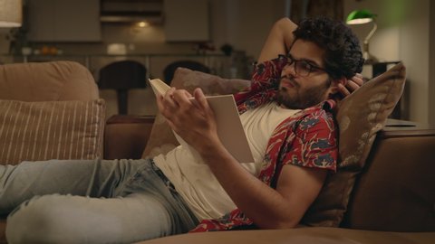 Young attractive modern Asian Indian man or male wearing eyeglasses sitting on a couch comfortably spending leisure or free time by reading a book relaxing in an interior home setup in the night. 