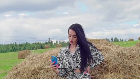 Sexy Brunette woman resting near hay in rural areas. Attractive model looking in phone camera device, make selfie picture