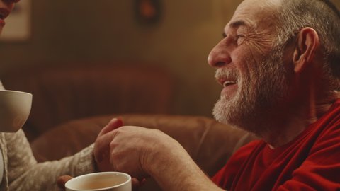 Elderly man kissing hand of happy woman and paying compliment while relaxing on couch and enjoying fresh tea in weekend at home