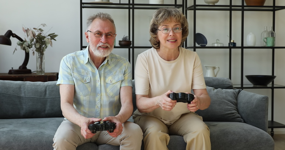 Happy elderly couple hold gamepads play arcade racing games sit on sofa have fun together at home, carefree 60s spouses enjoy competition spend free time use modern tech, digital entertainment concept Royalty-Free Stock Footage #1088175989