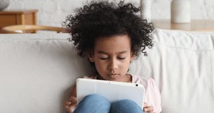 Adorable 5s African American kid girl spend time with gadget play online game watch video on electronic tablet alone. Little gen z child addicted to online activity use app on modern touchpad computer
