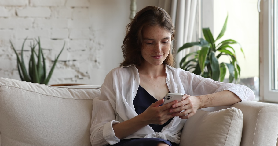 Pretty teen female sit on sofa at living room messaging with boyfriend in chat networking at social media using phone. Smiling young lady text answer on email play web game using wifi on smartphone Royalty-Free Stock Footage #1088176089