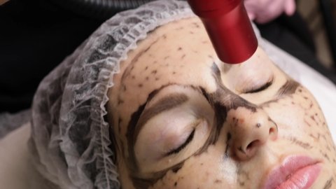 beautician applies black mask on face of beautiful woman. gorgeous woman in spa salon doing carbon peeling procedures for face.