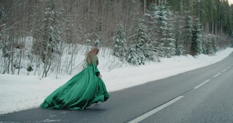 Caucasian blonde woman long hair walking running on winter snowing asphalt road in green dress brutal shoes. Fantasy girl runs winter forest. long vintage dress fabric waving. Slow motion 120 fps 4k