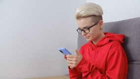 Tom boy female using mobile phone for communication and entertainment online. Stylish young woman with dyed and trimmed short hair browsing modern smartphone while sitting at home on couch 