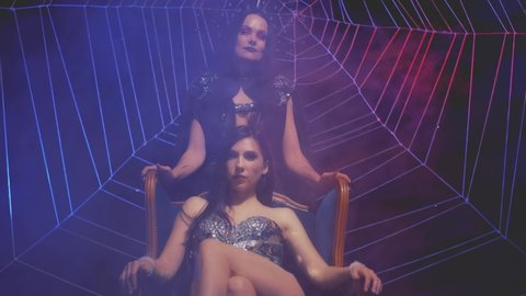 Two gothic fantasy women, dark queen of damned and black widow evil witch. Girl vampire in halloween costume. Woman princess sitting on throne. Background dark room spider web. Womens power concept