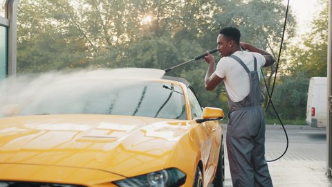 Handsome African guy in t-shirt and protective overalls washing his yellow luxury sport car, with high pressure water pump. Manual car wash with pressurized water in car wash outside. Car wash
