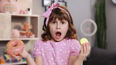 Little girl in pink dress holding donut and macaron in hands. Cute adolescent school girl plays with sweet donut macaroon doing happy fun face expressions on background. Funny concept with sweets.