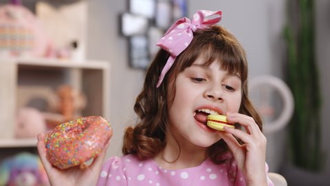 Adorable kid girl biting piece of donut macaron while sitting at table at home. Caucasian child school girl eating donut and yellow macaron and looks at camera. Sweet addiction concept