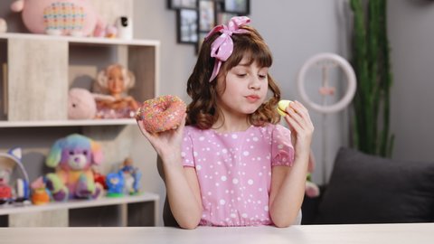 Caucasian child school girl eating donut and macaron at the same time. Adorable kid girl biting piece of donut macaron while sitting at table at home. Sweet addiction concept