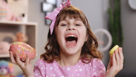 Adolescent school girl plays with sweet donuts doing happy fun face expressions on background. Cute little girl in pink dress holding macaron and donut in hands by the face. Funny concept with sweets