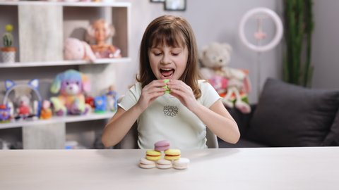 Happy smiling face of a little girl plays with dessert macarons on home background. Brunette kid girl bites pistachio green macaron cookie and looking at camera. Dessert person, sweet tooth, gourmet