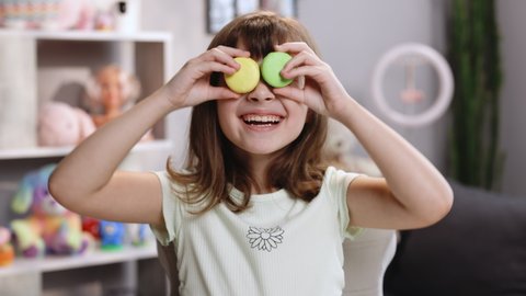 Teen girl plays with dessert macarons, holding the cookies like glasses around the eyes. Happy smiling face of a little girl covering her eyes with macaroons on a colored home background. Being happy