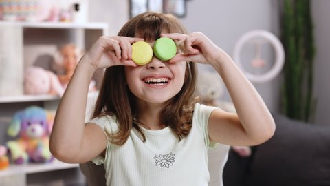 Teen girl plays with dessert macarons, holding the cookies like glasses around the eyes and throws his hands to the sides. Happy smiling face of little girl covering her eyes with macaroons
