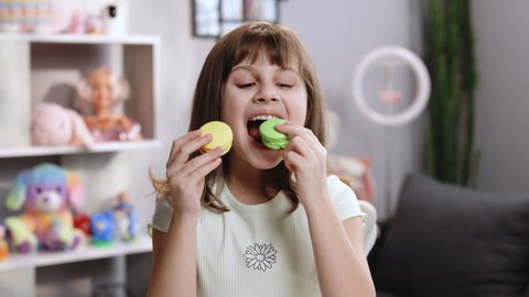 Attractive smiling cute preschool kid girl holds two tasty cookies in her hands and makes her choice, then bite macaron in green color. Traditional French multicolored macaroon. Food concept.