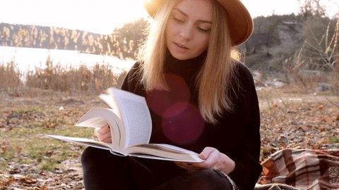 4k Caucasian blonde woman with beige hat in black sweater reads book in the countryside. Golden hour, cottagecore. Local travel. Slow living. Mental health