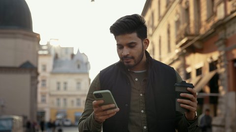 Handsome young guy feeling happiness while reading some good news on smartphone. Indian man in casual clothes standing on city street with cup of coffee.