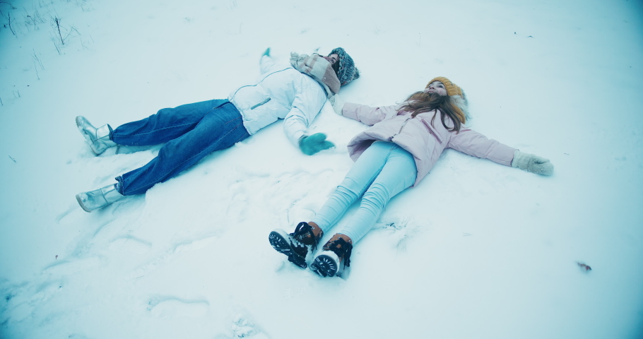 Woman and daughter lie on snowy ground making snow angels. Mother and daughter enjoy fresh air in forest on nasty cold winter day. Slow motion