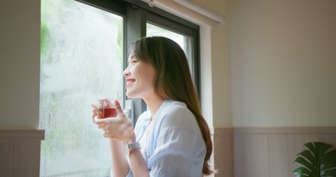 asian young woman is drinking tea or coffee while she looking through window in a rainy day at home
