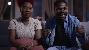 Young couple watching sports match and cheering for team, sitting on couch at home spbas. 4k video Close-up view of beautiful african woman, man watch tv and cheer with smiles, have fun and sit on